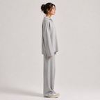 A woman stands in profile wearing a light gray knit lounge set consisting of a long-sleeve top and matching wide-leg pants, paired with white sneakers, against a plain light gray studio background.