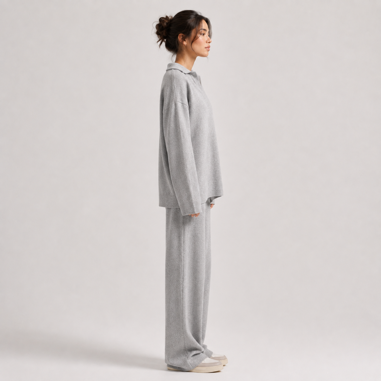 A woman stands in profile wearing a light gray knit lounge set consisting of a long-sleeve top and matching wide-leg pants, paired with white sneakers, against a plain light gray studio background.