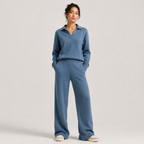 A woman modeling a matching knit loungewear set consisting of a long-sleeve polo-neck sweater and wide-leg pants in a dusty blue color, paired with white sneakers, against a light gray studio background.