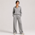 A woman modeling a light gray knit matching loungewear set consisting of a long-sleeve polo-neck top and wide-leg pants, standing against a neutral light gray studio background.
