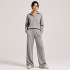 A woman modeling a light gray knit matching loungewear set consisting of a long-sleeve polo-neck top and wide-leg pants, standing against a neutral light gray studio background.