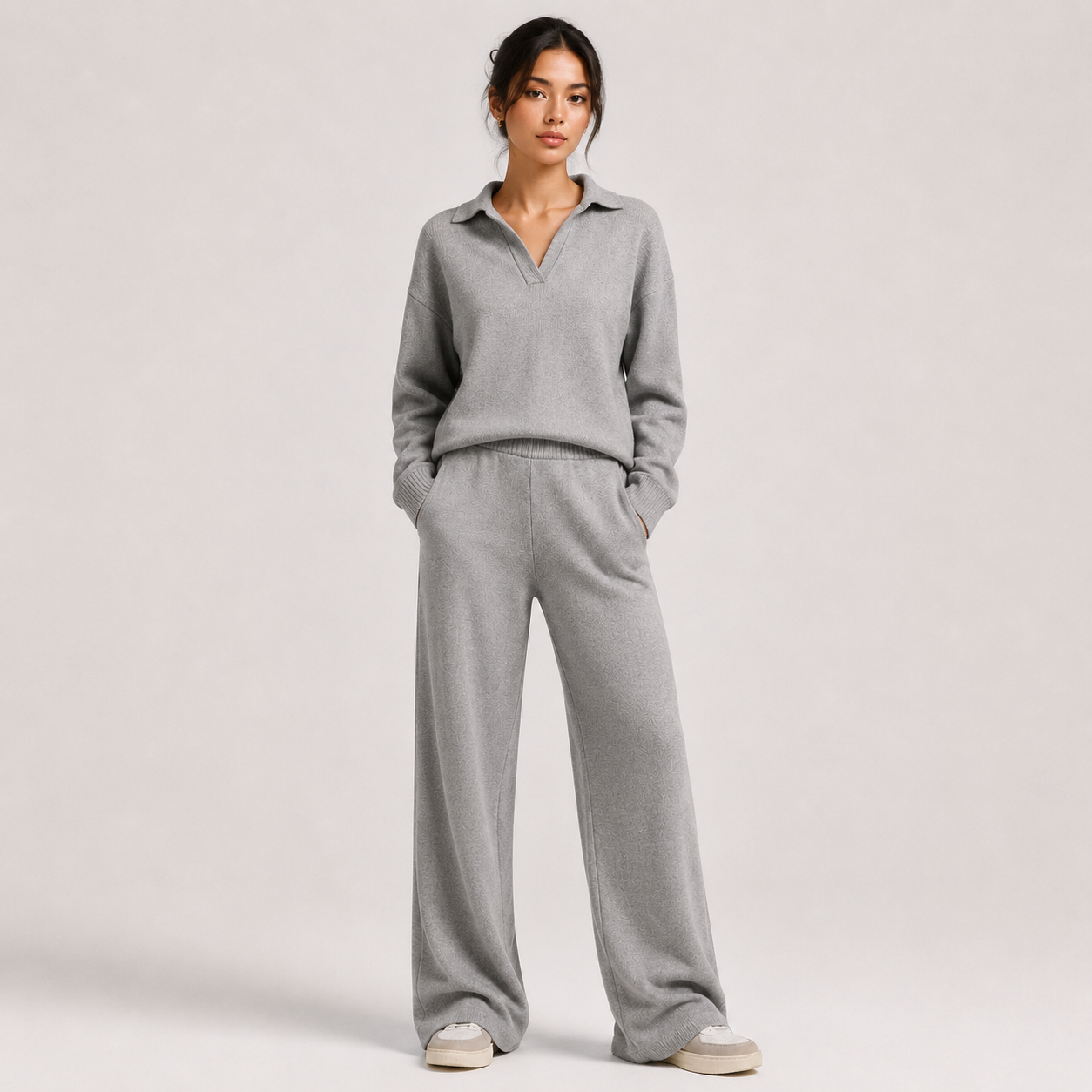 A woman modeling a light gray knit matching loungewear set consisting of a long-sleeve polo-neck top and wide-leg pants, standing against a neutral light gray studio background.