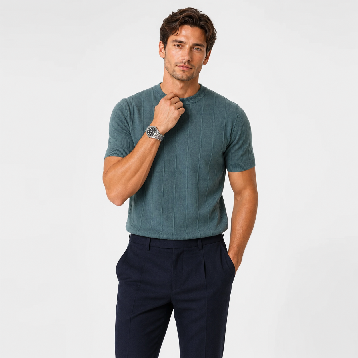 A man wearing a teal ribbed short-sleeve knit crewneck sweater and navy trousers, standing against a light gray studio background.