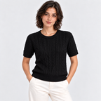 A woman wearing a black short-sleeve cable-knit crewneck sweater and off-white trousers, standing against a light gray studio background.