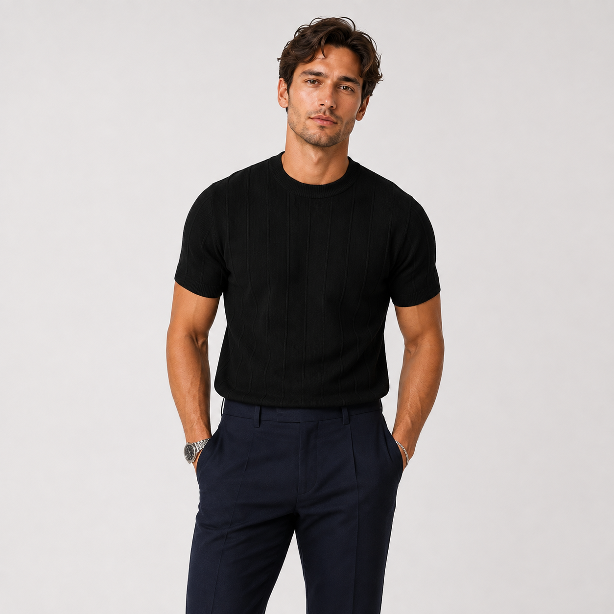 A man wearing a black short-sleeve ribbed knit crewneck t-shirt and navy blue trousers, standing against a light gray studio background.