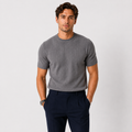 A man wearing a gray short-sleeve ribbed knit crewneck t-shirt and navy blue trousers, standing against a light gray studio background.