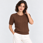 A woman wearing a brown short-sleeve cable-knit sweater and white pants, standing against a light gray studio background.