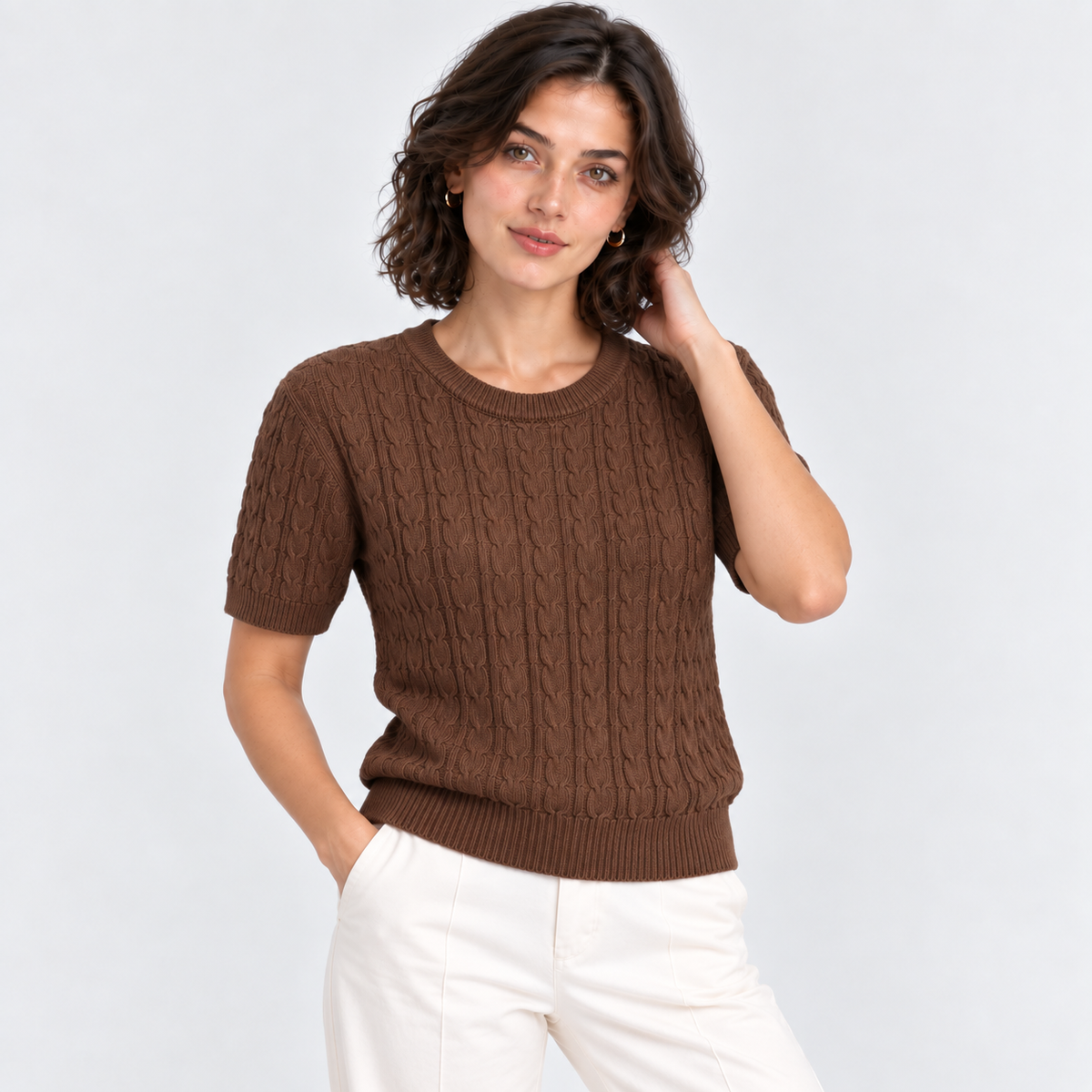A woman wearing a brown short-sleeve cable-knit sweater and white pants, standing against a light gray studio background.