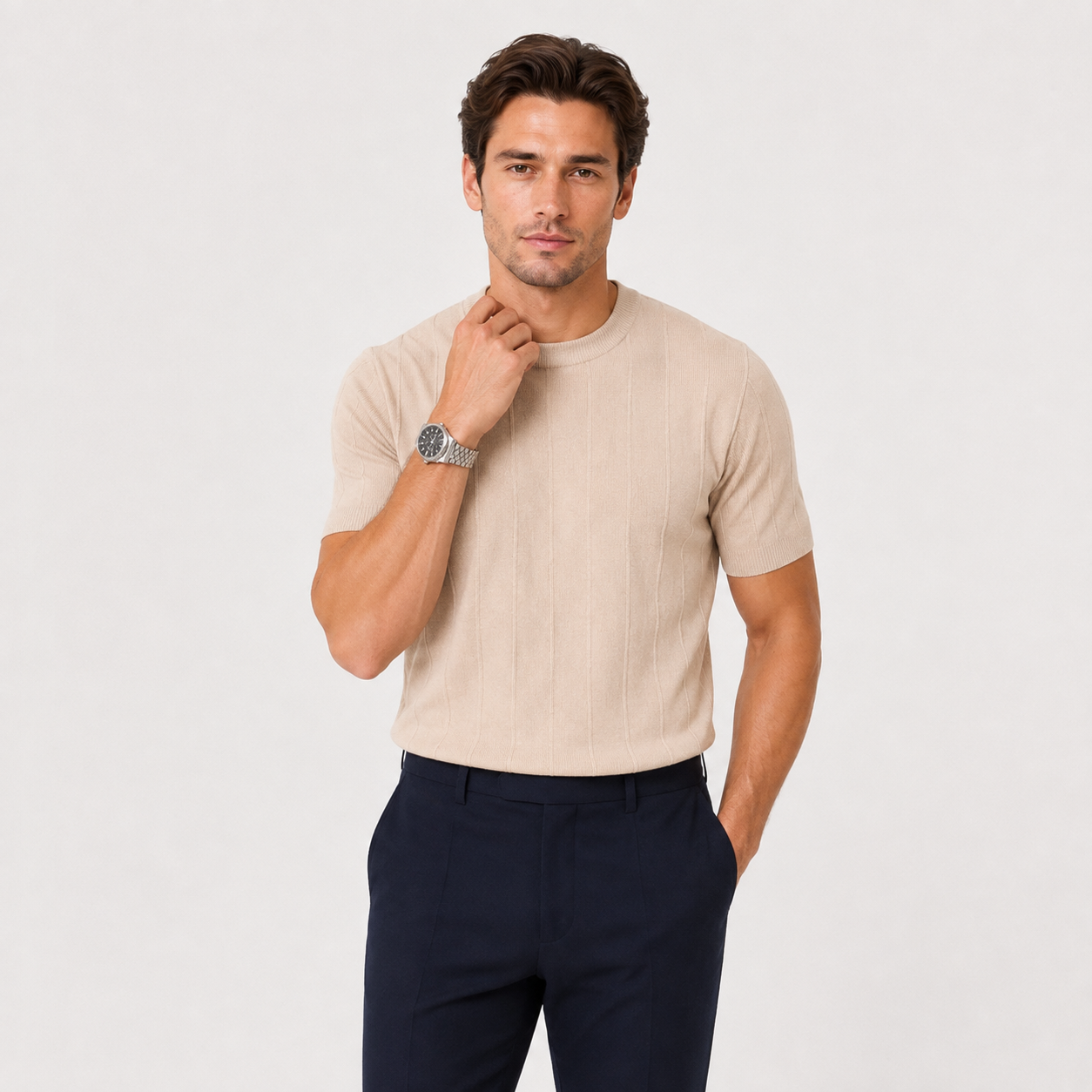 A man wearing a beige short-sleeve ribbed knit crewneck t-shirt and navy blue trousers, standing against a light gray studio background.