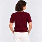 A woman shown from the back wearing a short-sleeve burgundy cable-knit sweater and white pants, photographed against a light gray studio background.