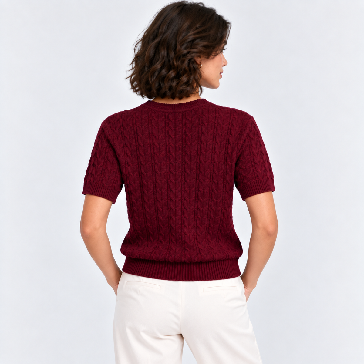 A woman shown from the back wearing a short-sleeve burgundy cable-knit sweater and white pants, photographed against a light gray studio background.
