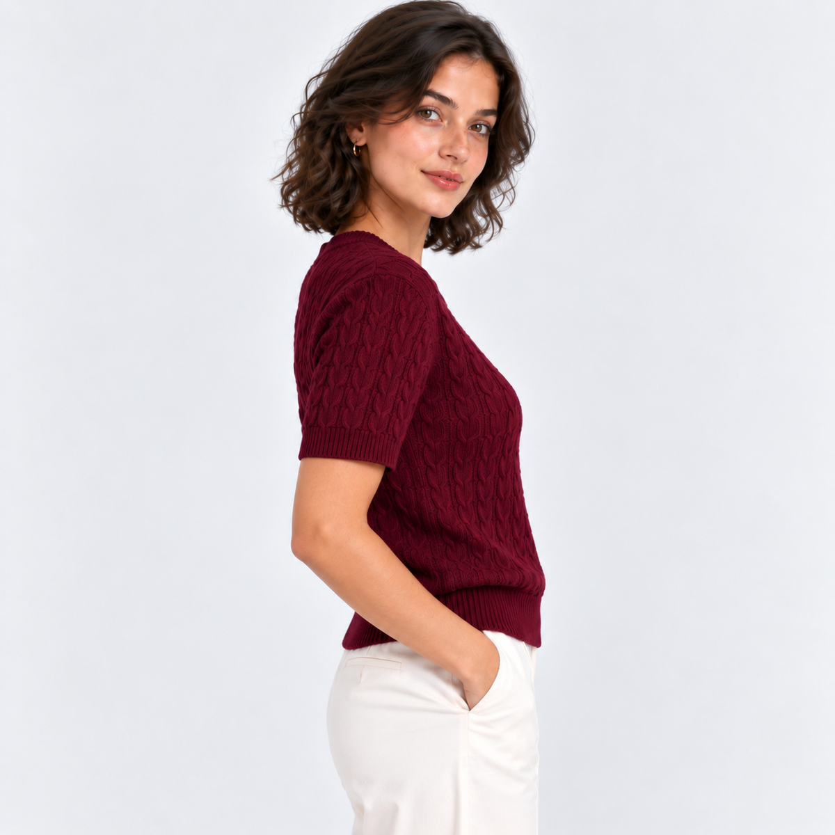 A woman wearing a short-sleeve burgundy cable-knit sweater and white trousers, shown from a side-back angle against a light gray studio background.