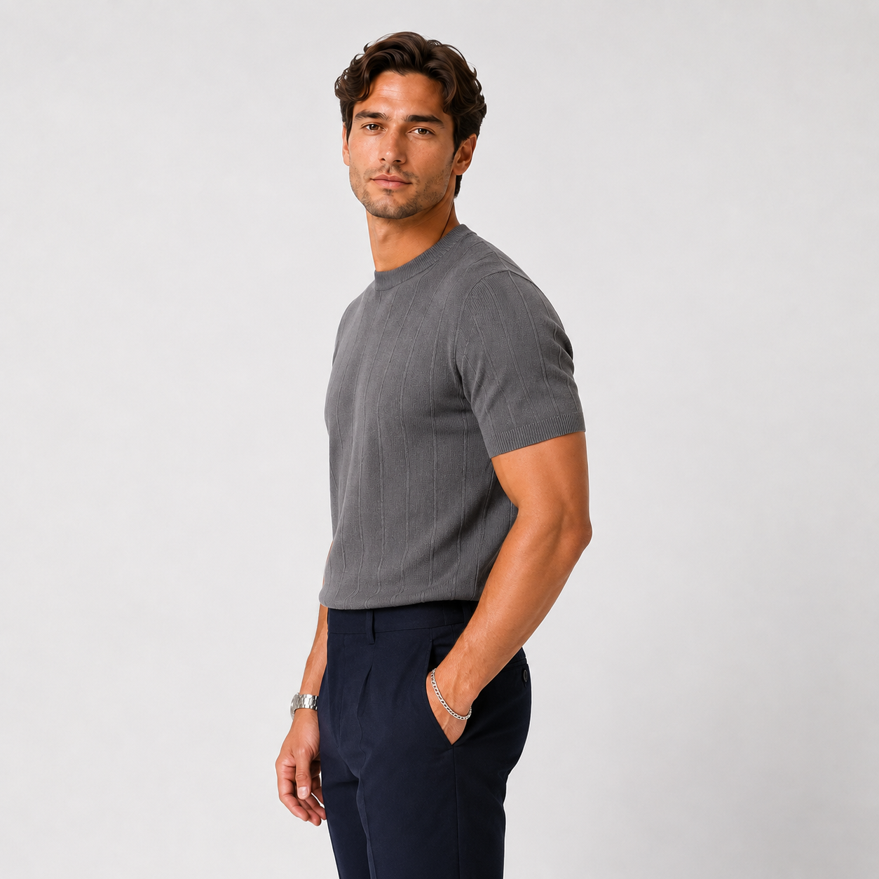 A man wearing a dark gray ribbed short-sleeve crewneck t-shirt and navy blue trousers, standing against a light gray studio background.