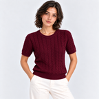 A woman wearing a short-sleeve burgundy cable-knit crewneck sweater and white trousers, standing against a light gray studio background.