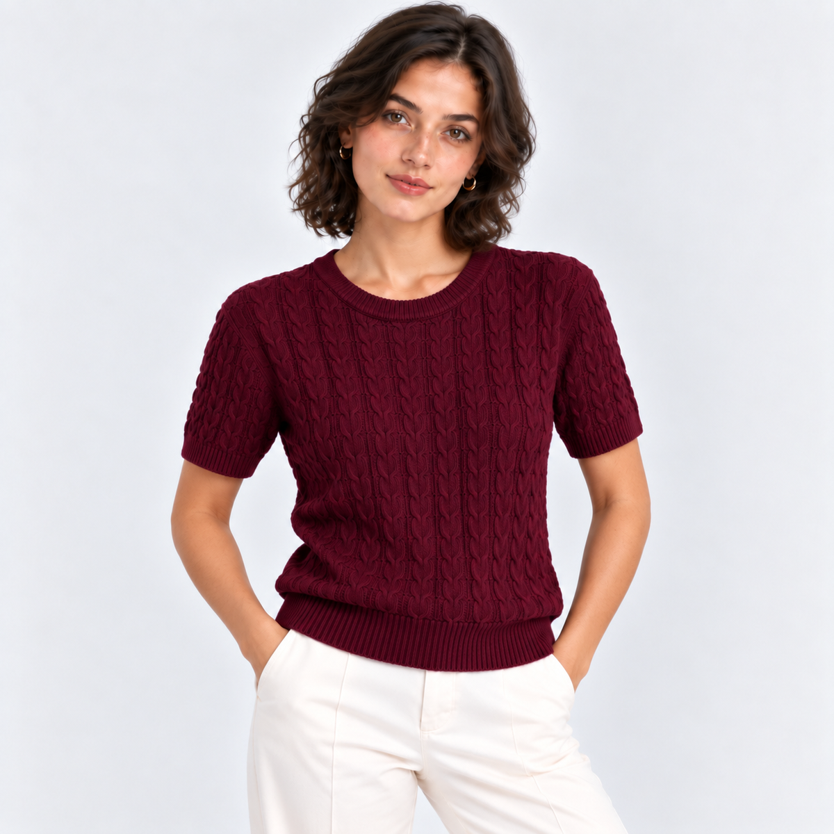 A woman wearing a short-sleeve burgundy cable-knit crewneck sweater and white trousers, standing against a light gray studio background.
