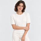 A woman modeling a short-sleeve crewneck cable-knit sweater in off-white, paired with matching off-white trousers, against a light gray studio background.