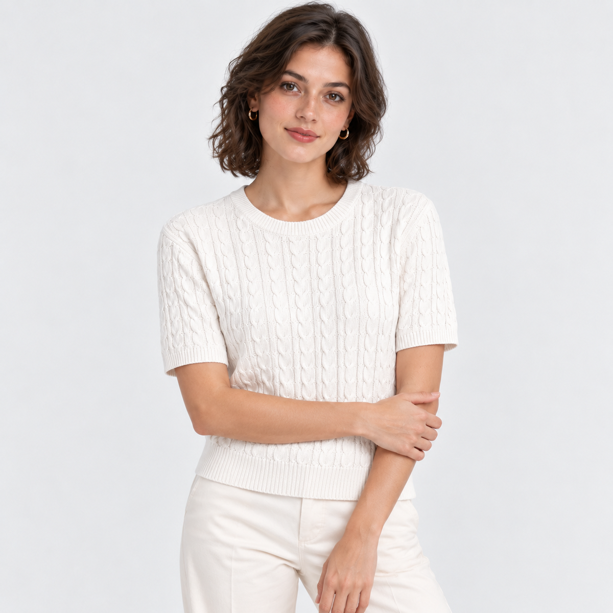 A woman modeling a short-sleeve crewneck cable-knit sweater in off-white, paired with matching off-white trousers, against a light gray studio background.