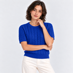 A woman wearing a bright royal blue short-sleeve cable-knit sweater and white pants, posing against a light gray studio background.