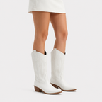 White western-style knee-high boots with brown heels, shown on a model wearing a white skirt against a light gray studio background.