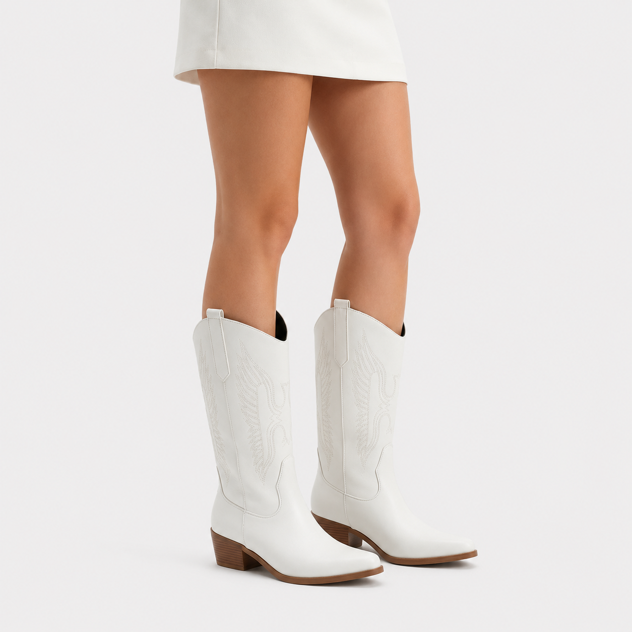 White western-style knee-high boots with brown heels, shown on a model wearing a white skirt against a light gray studio background.