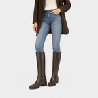 A model wearing blue skinny jeans, a brown coat, a cream sweater, and dark brown leather knee-high boots, shown from the waist down on a light gray background.