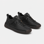 A pair of black low-top sneakers with a mix of leather and textured synthetic materials, featuring a hook-and-loop strap closure, laces, and a distinctive stitched midsole, shown on a light gray background.