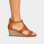 A woman's tan leather wedge sandal with an ankle strap and open toe, shown on a model's foot against a light gray background.