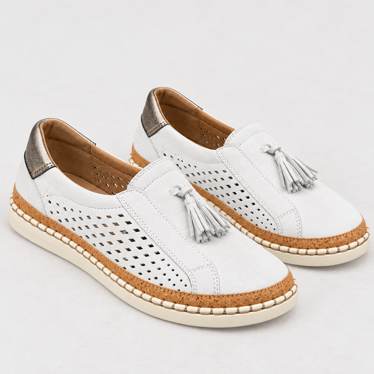 A pair of white slip-on espadrille-style shoes with perforated side panels, silver tassels, tan braided trim, and metallic silver heel tabs, shown on a light gray background.