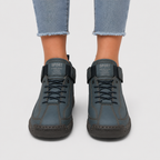 A pair of dark slate blue high-top sneakers with hook-and-loop strap closures and lace-up front, shown on a model wearing light-wash frayed-hem jeans against a light gray background.