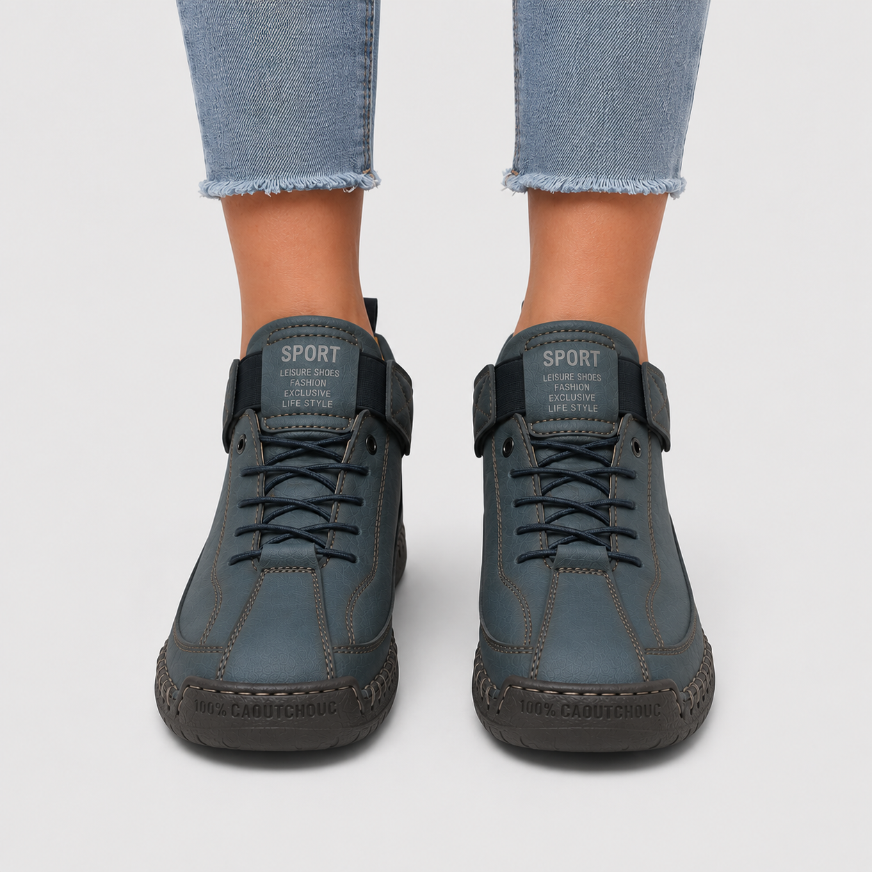 A pair of dark slate blue high-top sneakers with hook-and-loop strap closures and lace-up front, shown on a model wearing light-wash frayed-hem jeans against a light gray background.