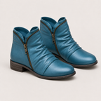 A pair of teal blue leather ankle boots with side zippers, low block heels, and dark brown soles, shown on a light beige background.