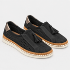 A pair of black slip-on espadrille-style shoes with perforated suede-like uppers, black tassels, cork-wrapped midsoles, and off-white soles, photographed on a light gray studio background.