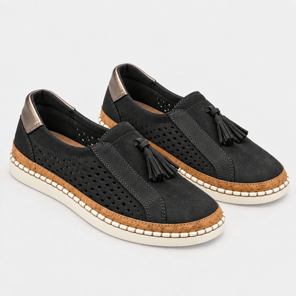 A pair of black slip-on espadrille-style shoes with perforated suede-like uppers, black tassels, cork-wrapped midsoles, and off-white soles, photographed on a light gray studio background.