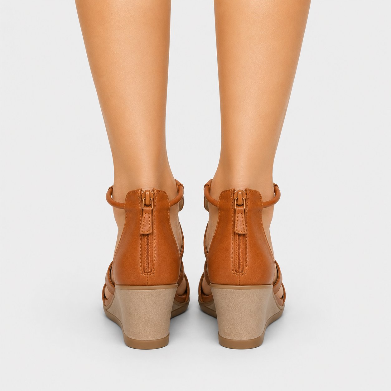 Rear view of a person wearing tan leather wedge sandals with ankle straps and low block heels on a light gray background.