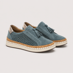 A pair of women's slip-on tassel loafers in dusty blue suede, featuring perforated side panels, a cork-wrapped midsole, and an off-white rubber outsole, shown against a light gray background.