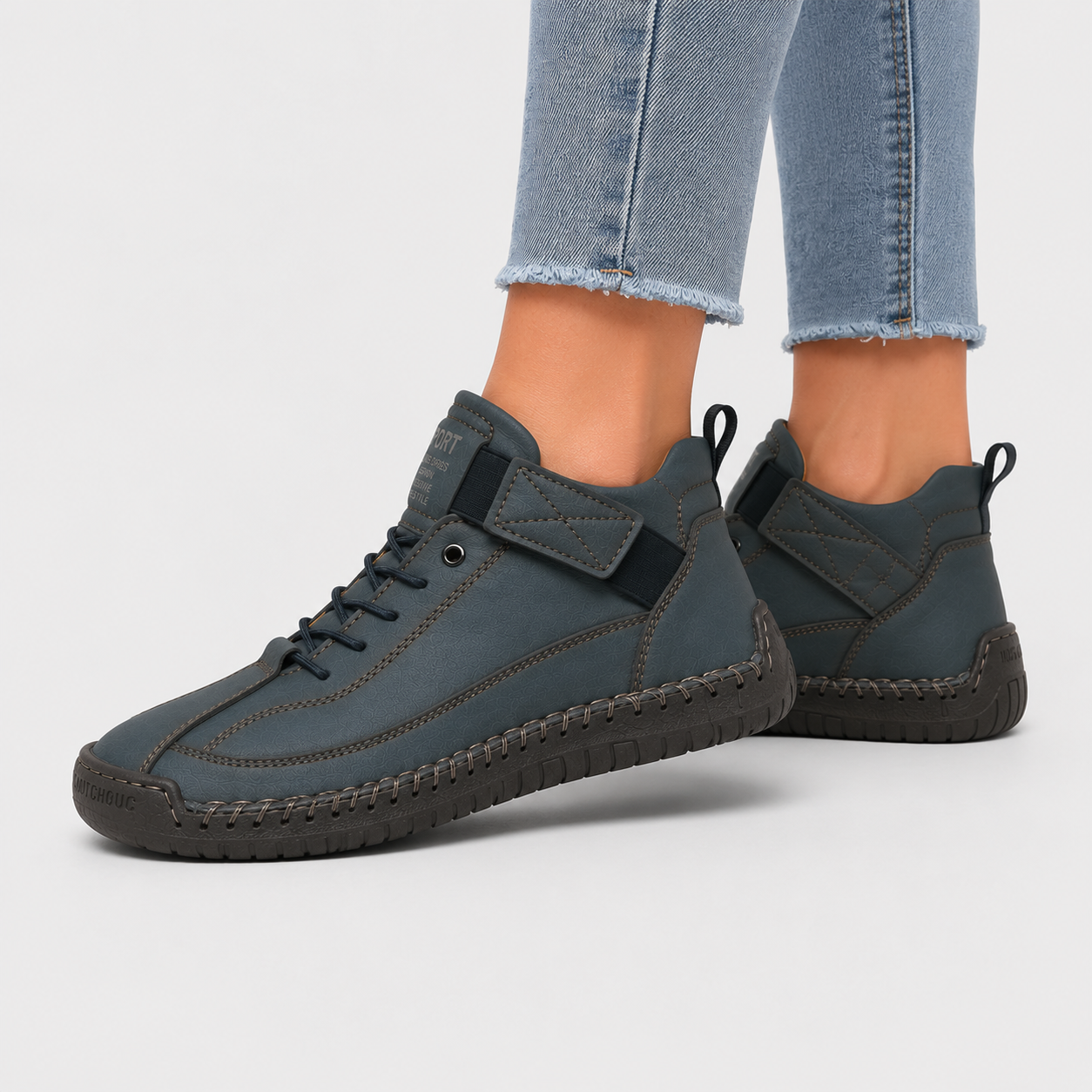 A pair of slate blue high-top sneakers with dark soles, shown on a model's feet wearing light-wash frayed-hem jeans against a light gray studio background.