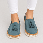 A pair of women's slip-on espadrille-style shoes in a muted teal blue suede-like material, featuring dark blue tassels, perforated sides, cork midsoles, and cream rubber outsoles. The shoes are shown on a model's feet wearing white pants against a light gray studio background.
