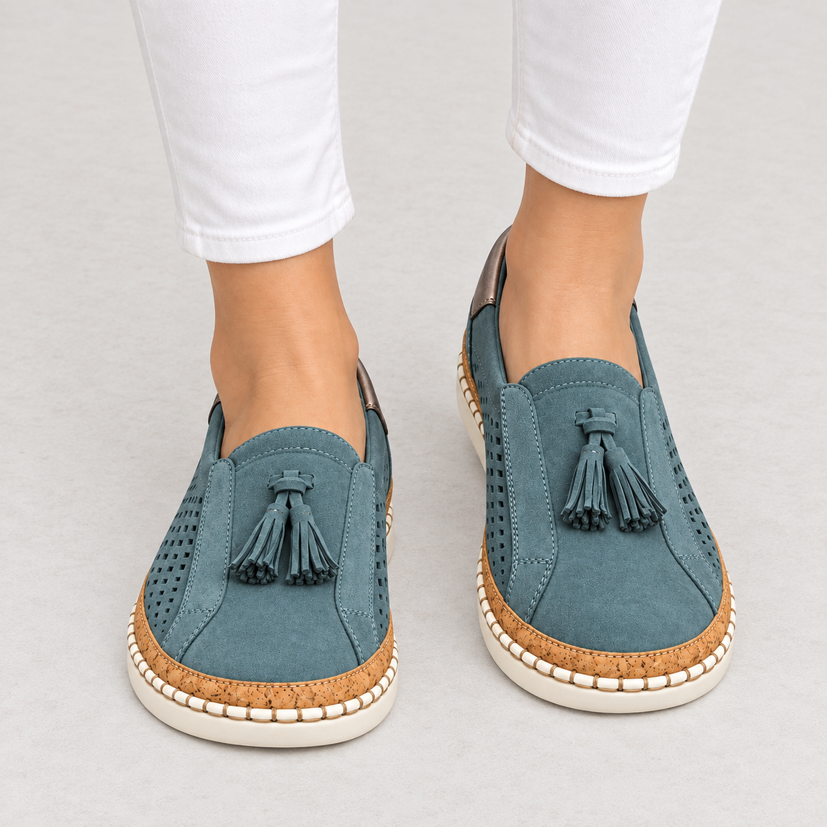 A pair of women's slip-on espadrille-style shoes in a muted teal blue suede-like material, featuring dark blue tassels, perforated sides, cork midsoles, and cream rubber outsoles. The shoes are shown on a model's feet wearing white pants against a light gray studio background.