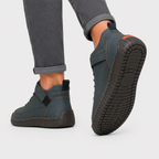 A pair of dark slate blue slip-on shoes with black soles, shown on a model wearing gray cuffed jeans against a light gray background.