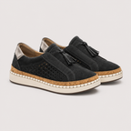 A pair of women's black suede slip-on shoes with tassel details, perforated side panels, a cork-wrapped midsole, and an off-white rubber outsole, shown against a light gray background.