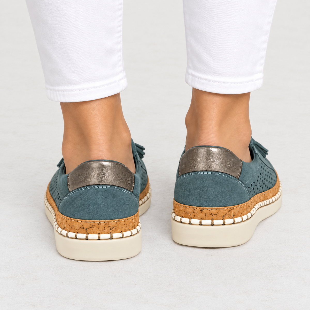 A pair of women's platform slip-on sneakers shown from the back, worn with white pants. The shoes feature a slate blue suede upper, a metallic silver heel tab, a cork midsole, and an off-white platform sole.