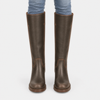 A pair of dark brown leather knee-high riding boots worn with blue jeans, shown from the back against a light gray background.