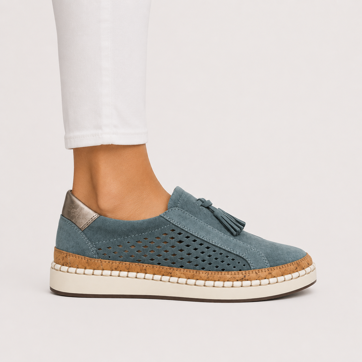 A blue suede slip-on platform sneaker with a tassel detail, side cutouts, and a cork-wrapped sole, shown on a model's foot wearing white pants against a light gray background.