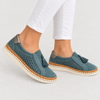 A pair of women's slip-on espadrille-style shoes in a muted teal blue suede-like material, featuring perforated sides, tassel details, cork-wrapped midsoles, and off-white soles. The shoes are shown on a model's feet wearing white pants against a light gray studio background.