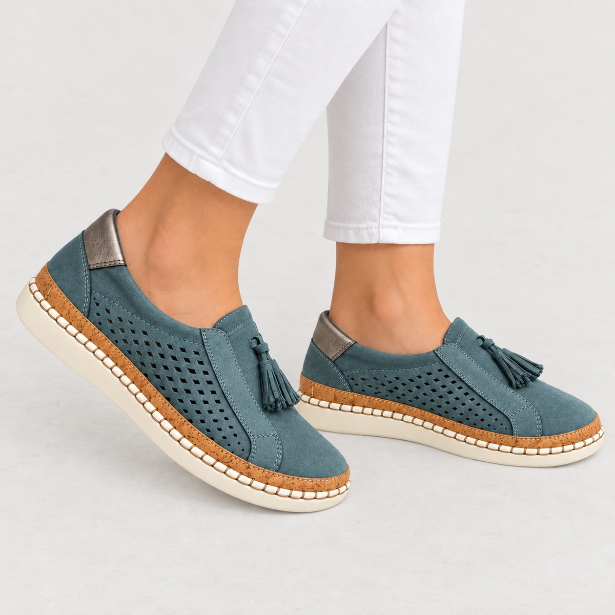 A pair of women's slip-on espadrille-style shoes in a muted teal blue suede-like material, featuring perforated sides, tassel details, cork-wrapped midsoles, and off-white soles. The shoes are shown on a model's feet wearing white pants against a light gray studio background.