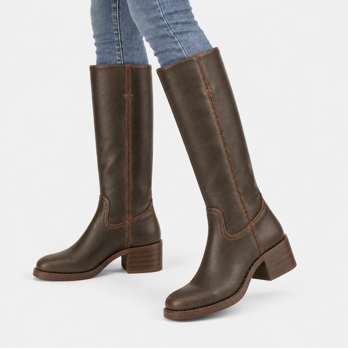 A pair of dark brown leather knee-high boots with low block heels, shown on a model wearing blue jeans against a light gray background.
