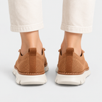 A pair of brown knit slip-on sneakers shown from the back, worn with off-white cropped pants against a clean light gray studio background.