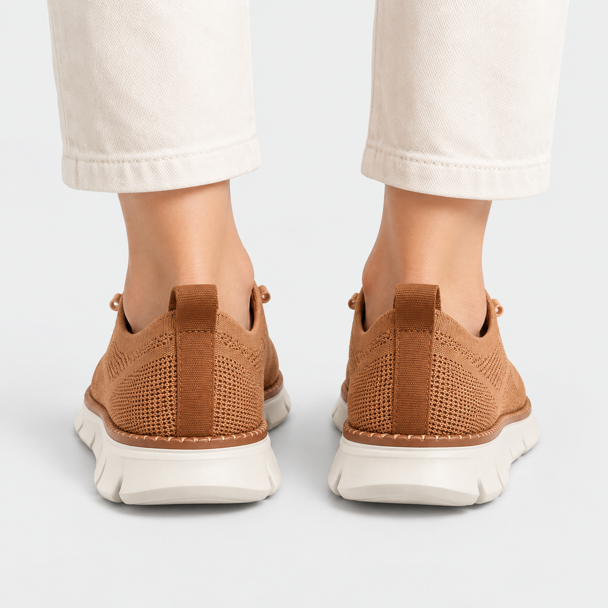A pair of brown knit slip-on sneakers shown from the back, worn with off-white cropped pants against a clean light gray studio background.