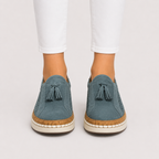 A pair of women's slip-on espadrille-style shoes in dusty blue suede with tassel details, worn with white pants against a light gray studio background.