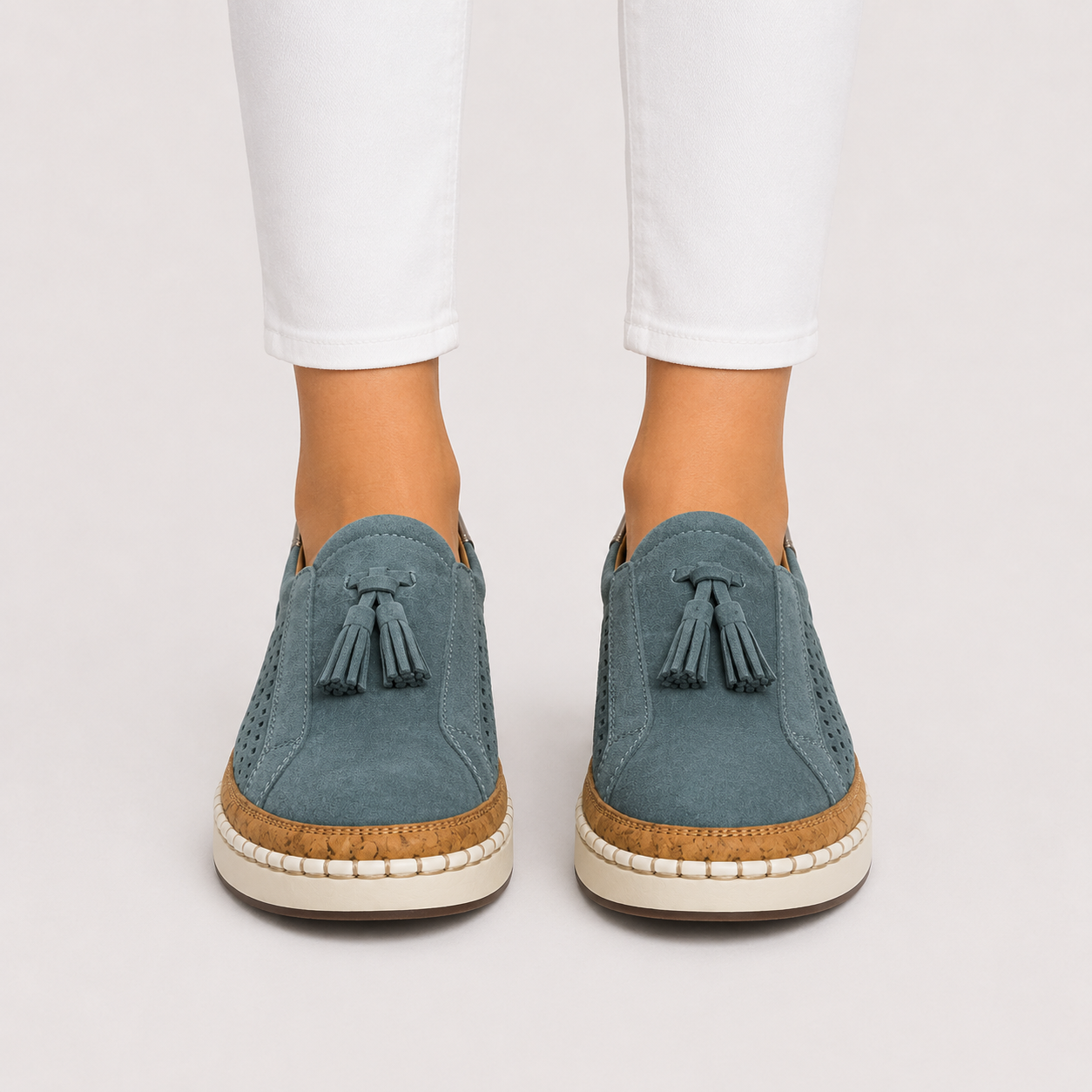 A pair of women's slip-on espadrille-style shoes in dusty blue suede with tassel details, worn with white pants against a light gray studio background.